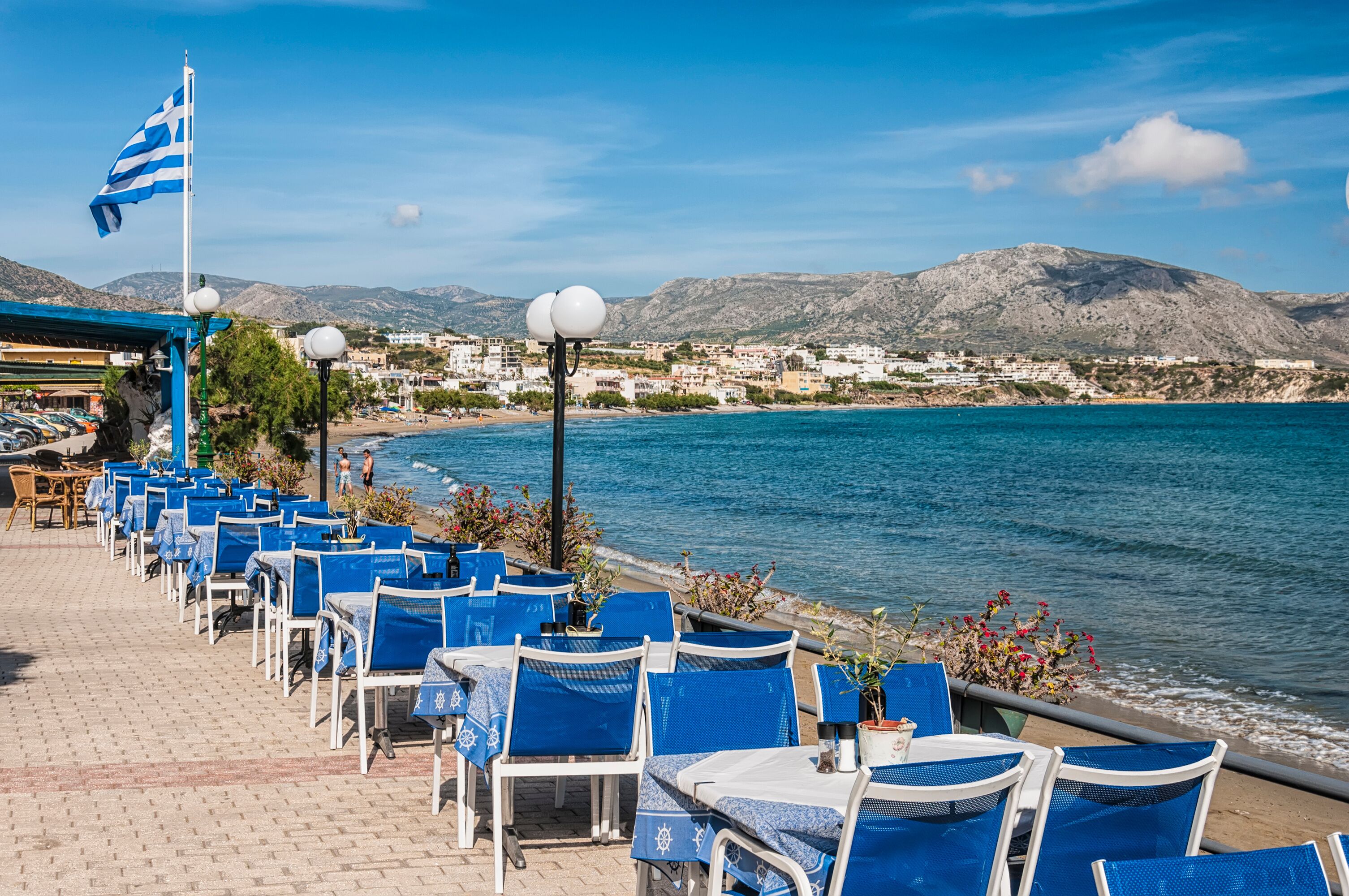 A seaside café with blue chairs and tables, a Greek flag, and a view of the sea and mountains.