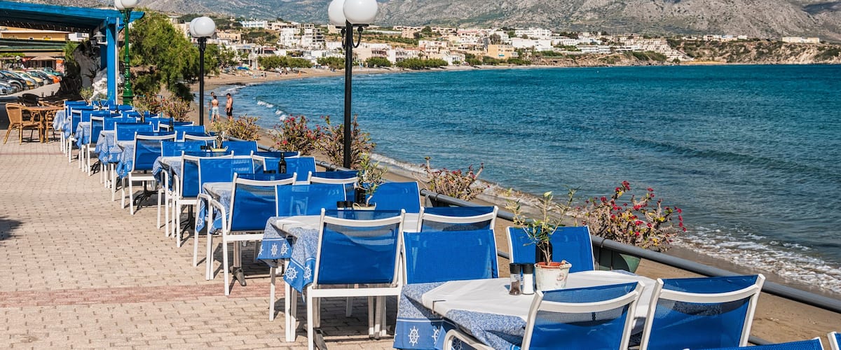 A seaside café with blue chairs and tables, a Greek flag, and a view of the sea and mountains.
