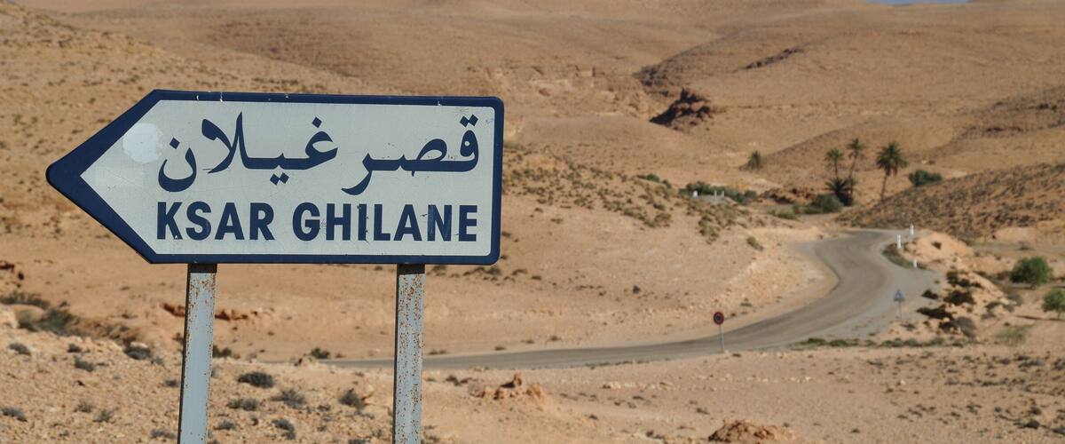 A desert landscape with a road curving through it and a sign pointing to 'KSAR GHILANE'.