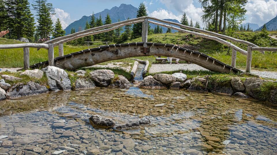 A wooden bridge over a clear stream with rocks, surrounded by mountains and trees.