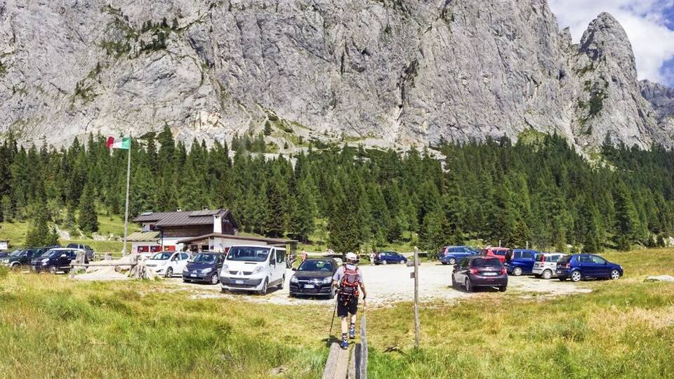 A mountainous landscape with a large rock formation, a parking area with several cars, and a small building.