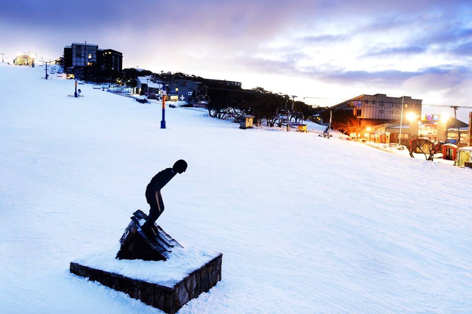 A snow-covered ski slope with a sculpture in the foreground, ski lifts, and buildings in the background.