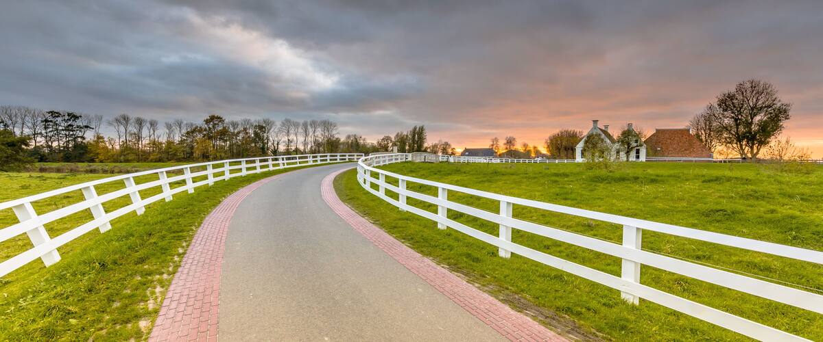 A curving white fence-lined path in a grassy field with a brick edge, leading towards a sunset over a rural landscape with trees and a small building.