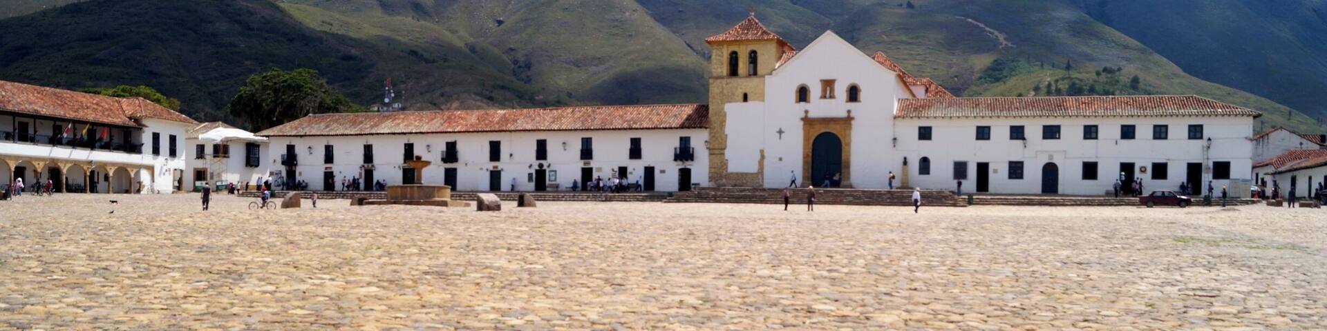 A cobblestone plaza with a white building with a red-tiled roof and a bell tower, surrounded by mountains.