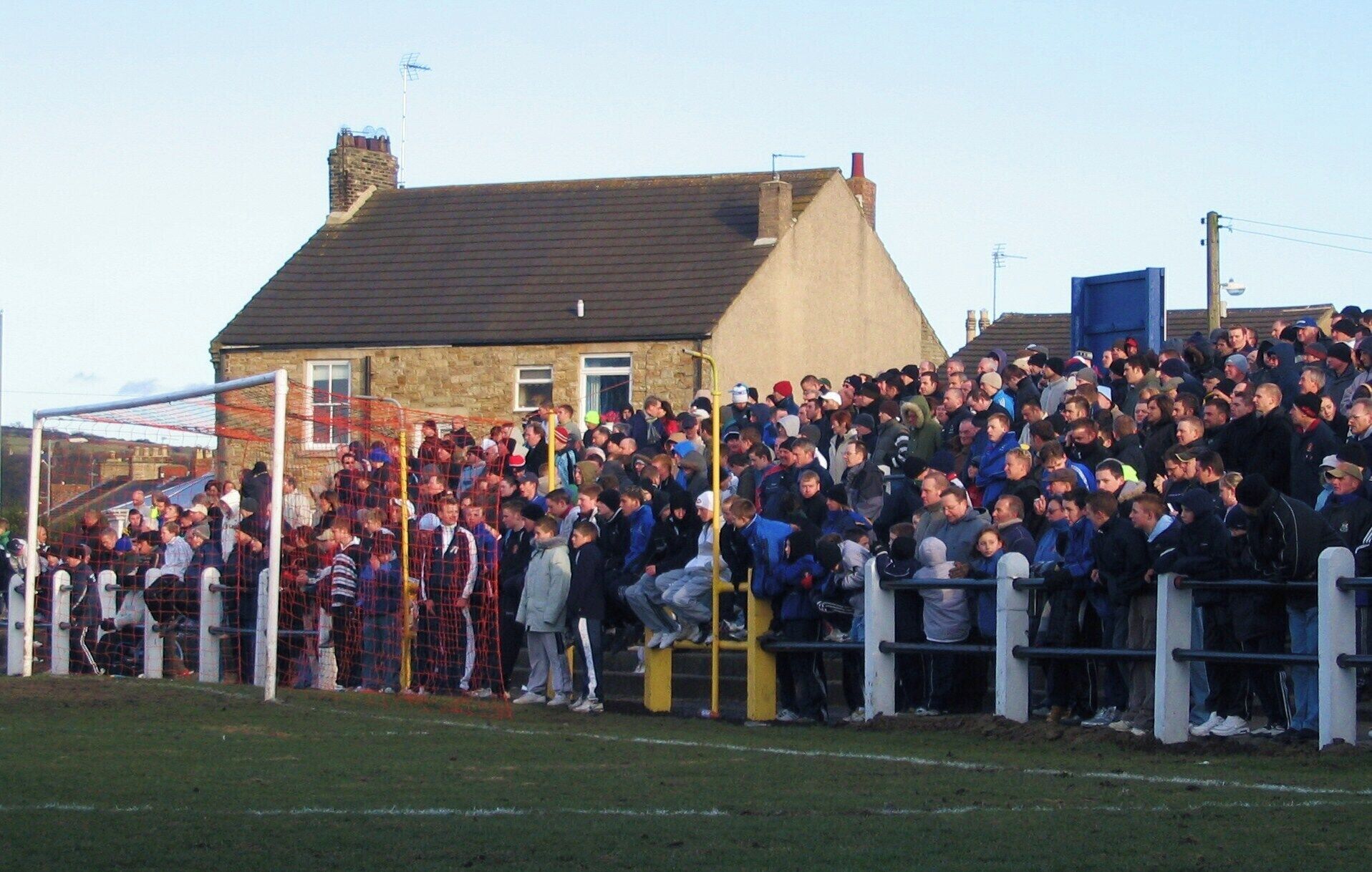 A large crowd of people gathered at a sports event, standing behind a fence with a goalpost visible in the foreground.