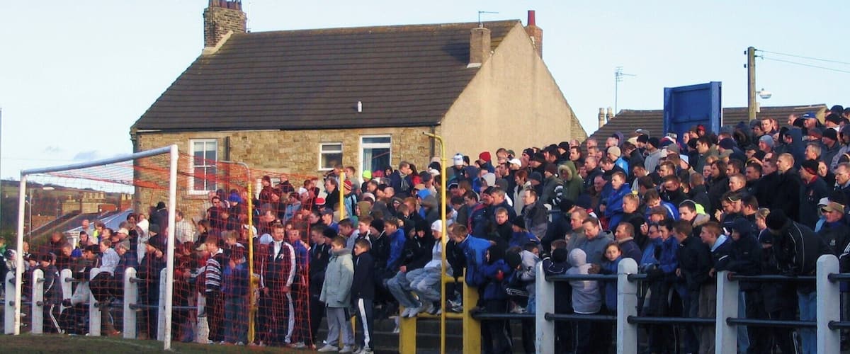 A large crowd of people gathered at a sports event, standing behind a fence with a goalpost visible in the foreground.