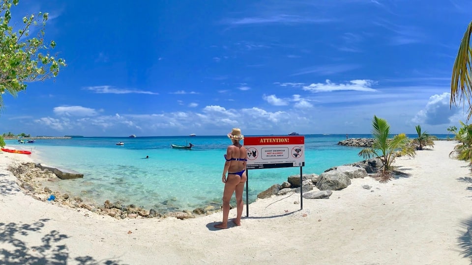 A beach with clear turquoise water, a sign warning about short-shore shark activity, and a person standing by the sign.