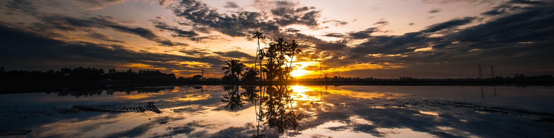A sunset over a calm lake with reflections of palm trees and a cloudy sky.