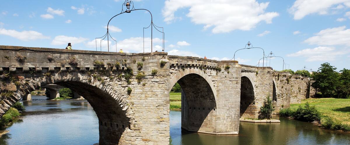 A stone bridge with multiple arches over a river, surrounded by greenery and clear skies.