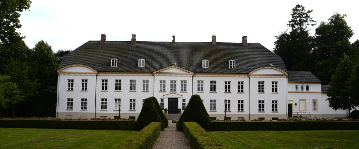A large white building with a symmetrical facade, a gravel pathway leading to the entrance, and well-manicured hedges.