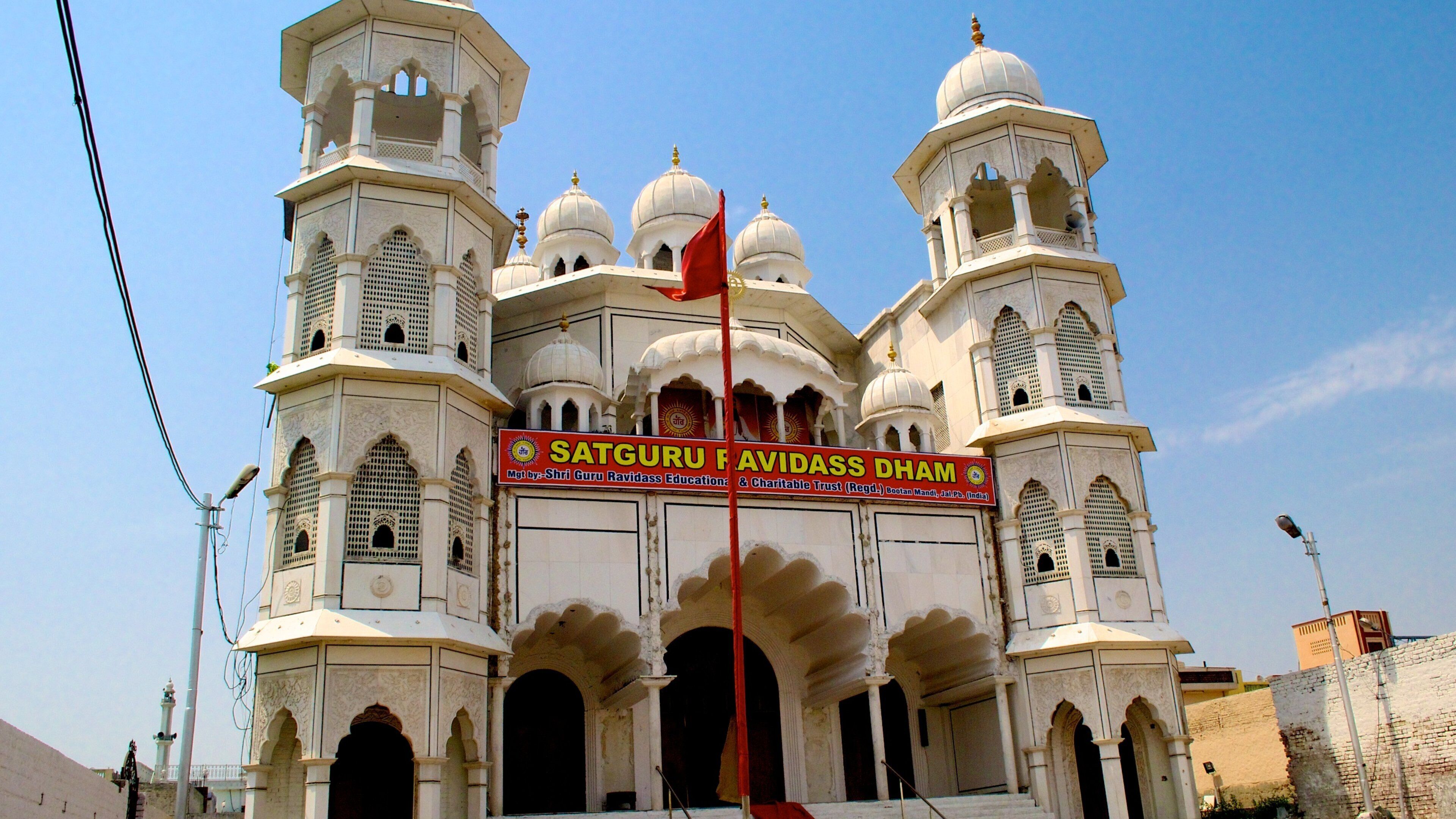 A large white building with multiple domes and minarets, featuring a prominent red flag and a sign reading "Satguru Ravidass Dhamm".