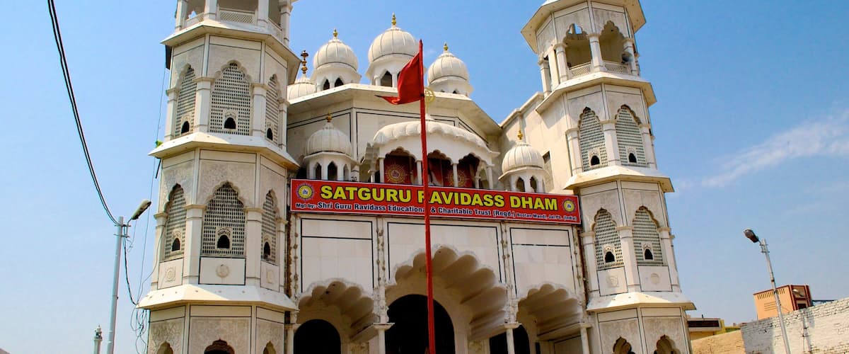 A large white building with multiple domes and minarets, featuring a prominent red flag and a sign reading "Satguru Ravidass Dhamm".