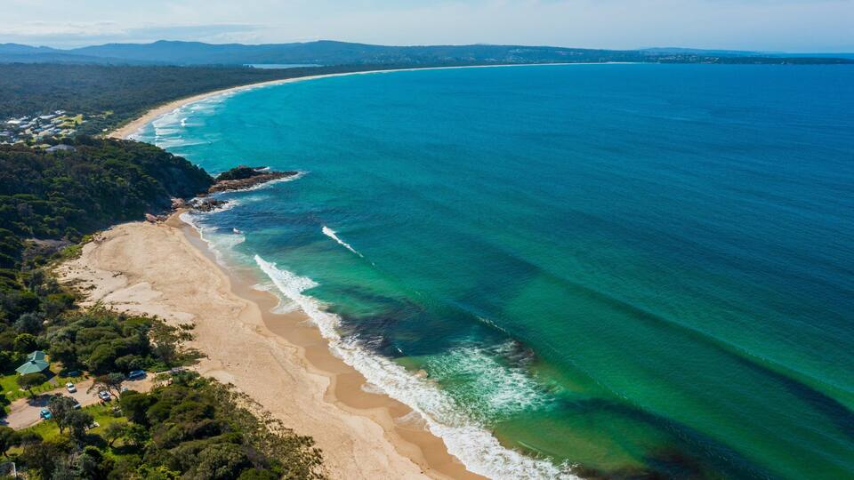 A coastal landscape with a sandy beach, clear turquoise waters, and a forested area.