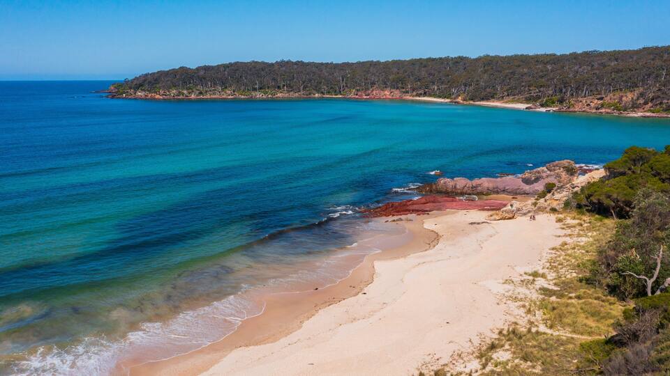 A coastal landscape with clear blue waters, a sandy beach, and a forested hill in the background.