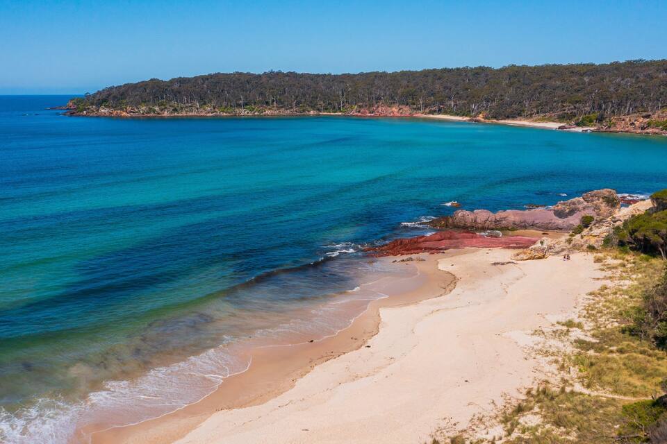 A coastal landscape with clear blue waters, a sandy beach, and a forested hill in the background.