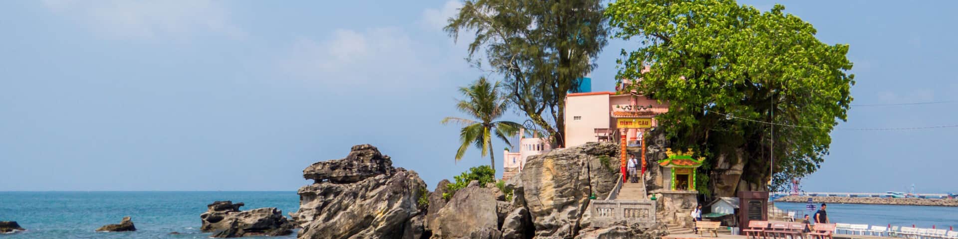A beach with clear turquoise water, rocky outcrops, a palm tree, and a building with a colorful facade.