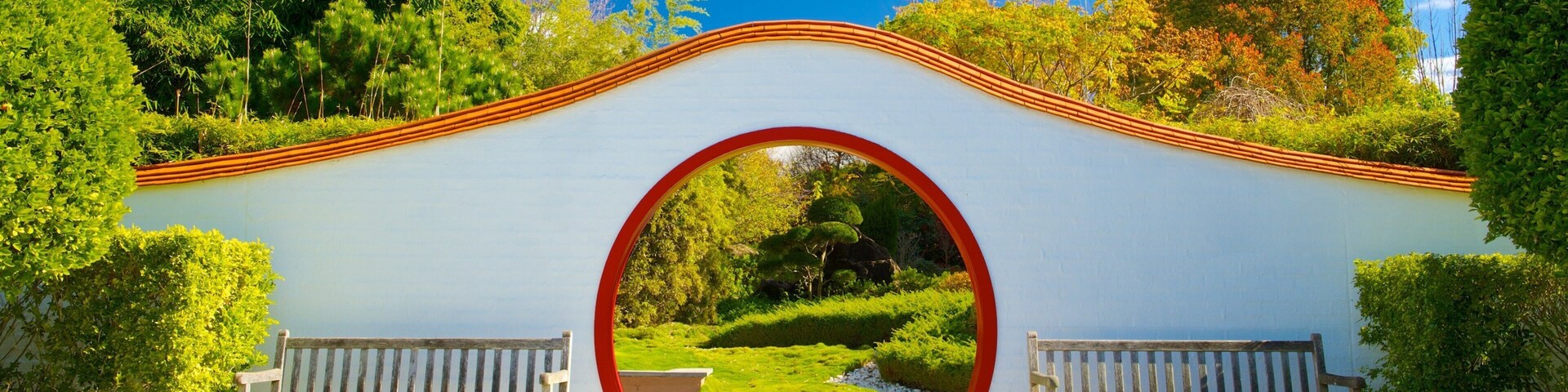 Uma trilha de pedra leva a um arco branco com um círculo vermelho no centro, ladeado por dois bancos de madeira.