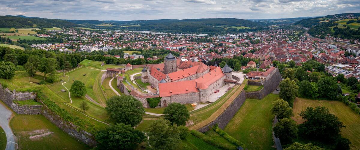 An aerial view of a historic town with a prominent castle surrounded by green hills and a river.