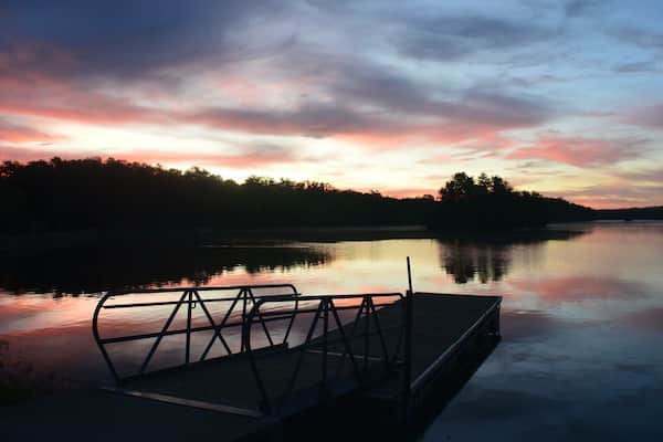 En rolig sø ved solnedgang med en dock og træer i baggrunden.
