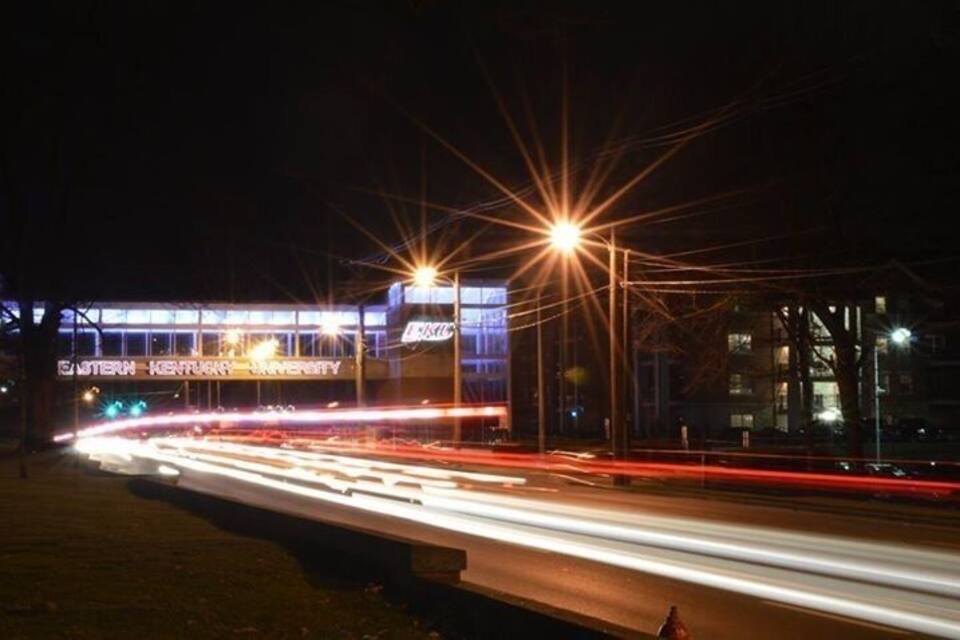 A nighttime cityscape with a lit-up building labeled "Eastern Kentucky University" and light trails from moving vehicles.