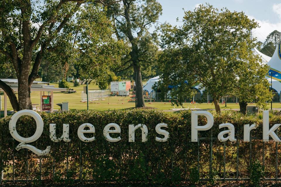 Placa indicando Queens Park, cercada por árvores e um campo esportivo.