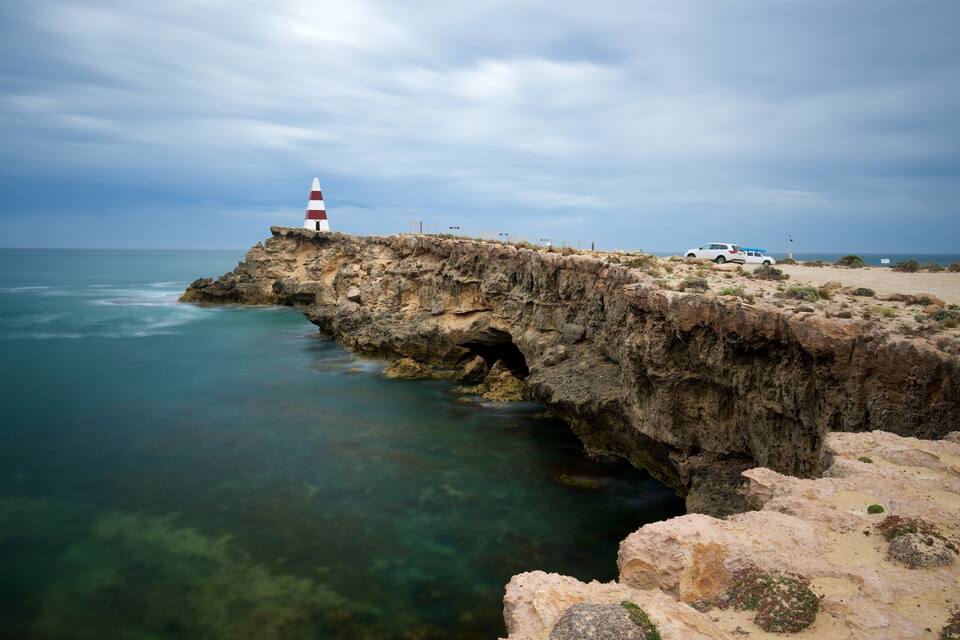 A rocky coastline with a lighthouse and a white car parked on the beach.