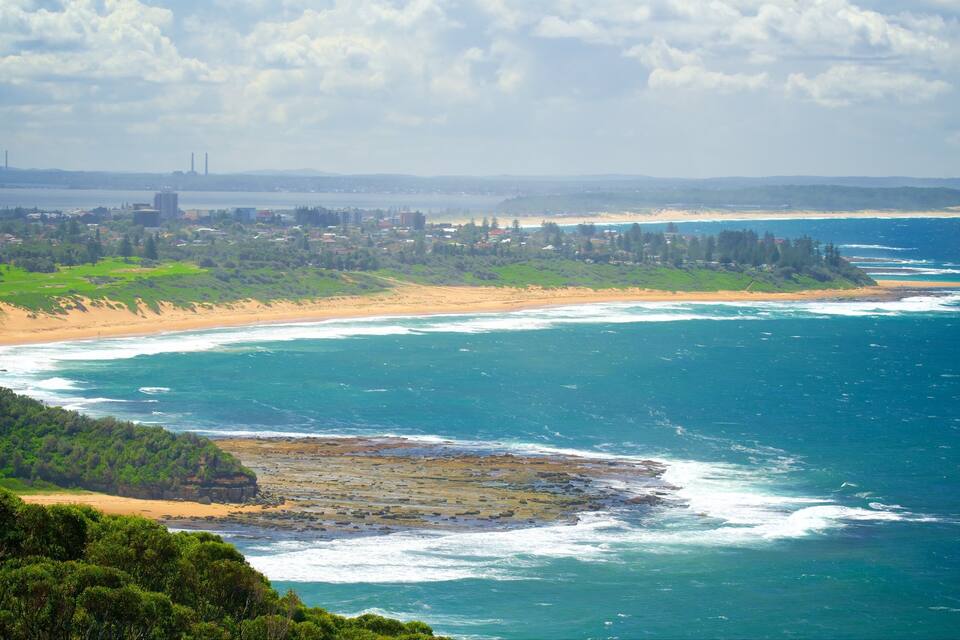 A coastal landscape with a sandy beach, ocean waves, and a distant city skyline.
