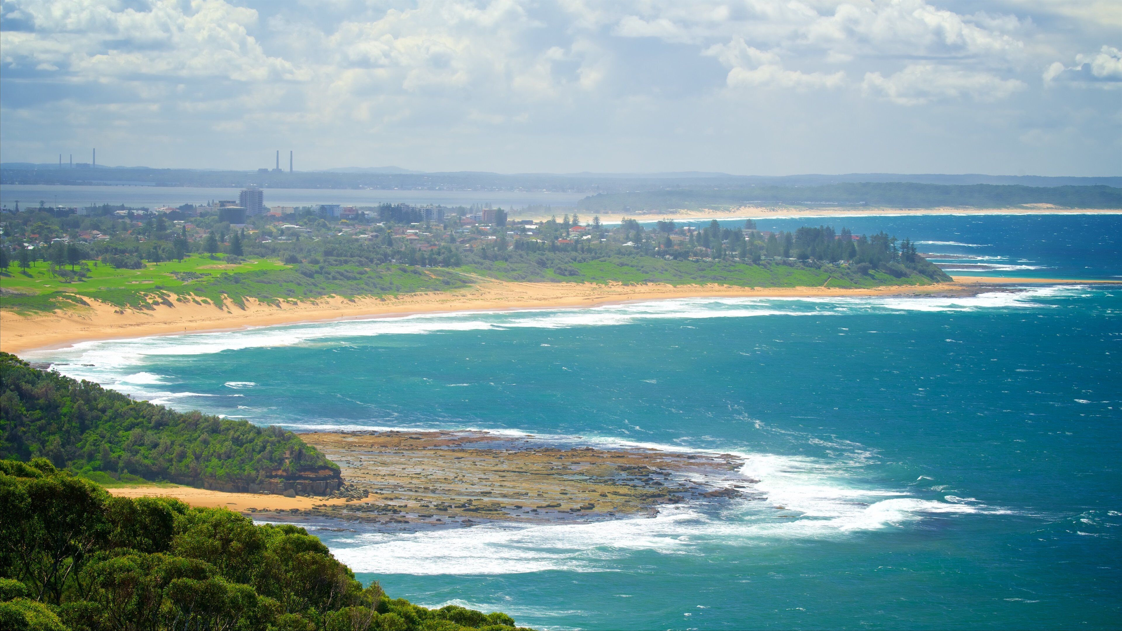 A coastal landscape with a sandy beach, ocean waves, and a distant city skyline.