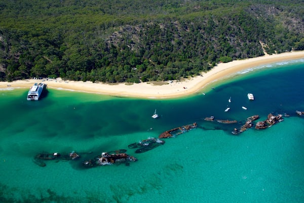 An aerial view of a coastal area with a sandy beach, clear turquoise waters, and several boats.