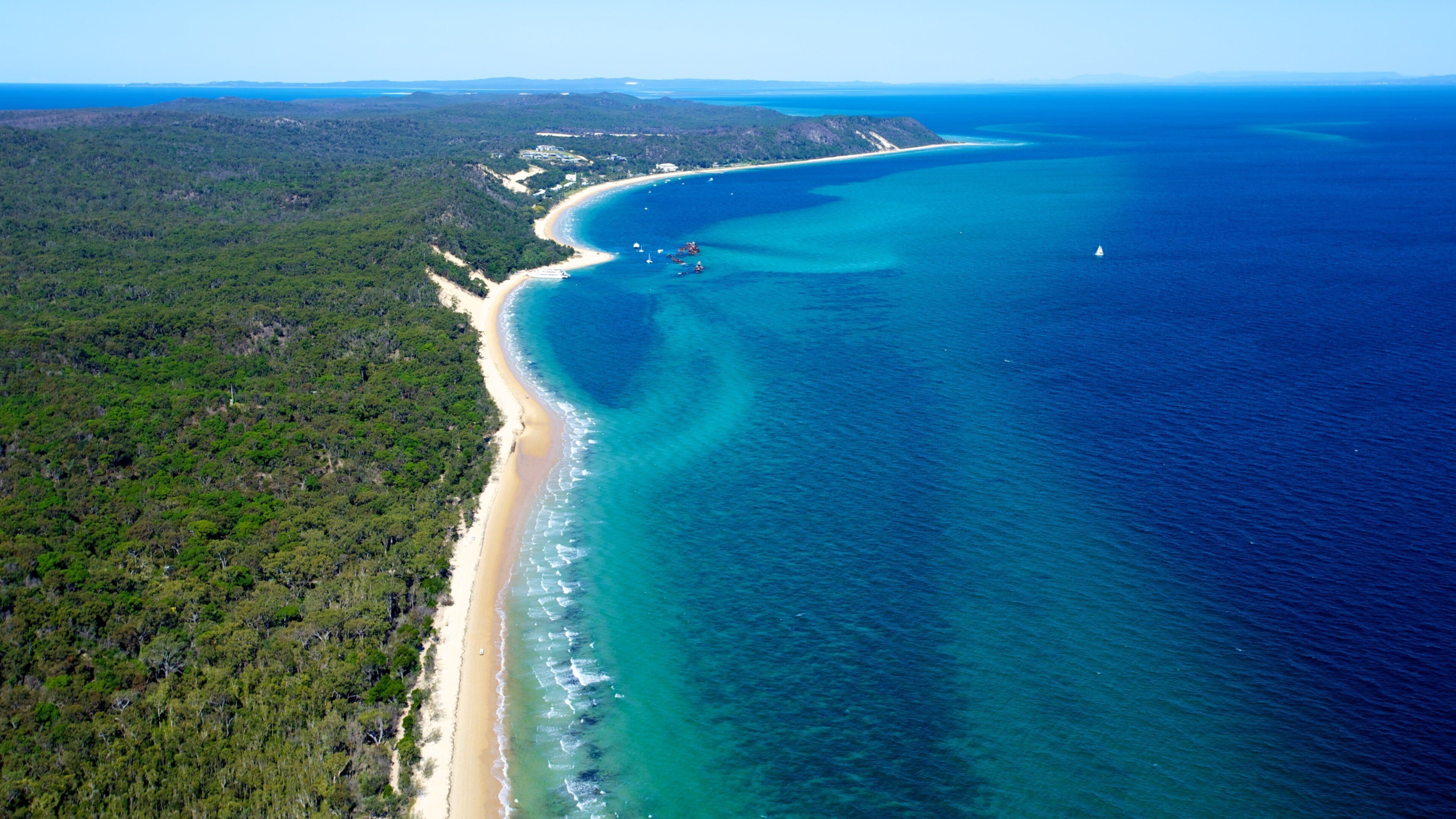 An aerial view of a coastal area with a sandy beach, clear blue waters, and dense green forest.