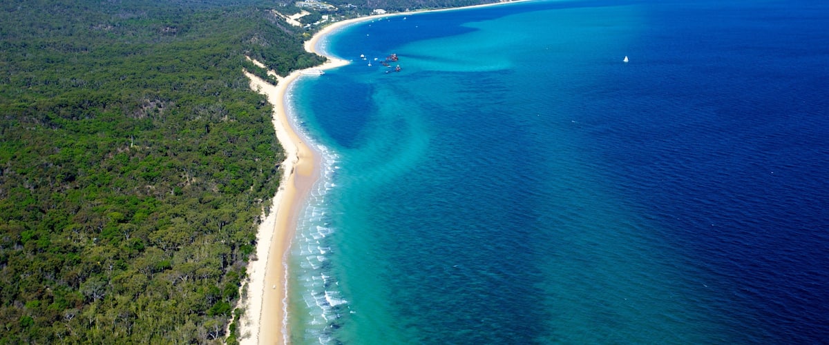 An aerial view of a coastal area with a sandy beach, clear blue waters, and dense green forest.