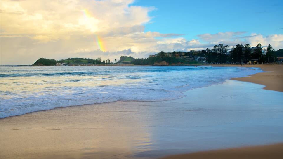 A beach with waves, a rainbow, and buildings in the background.