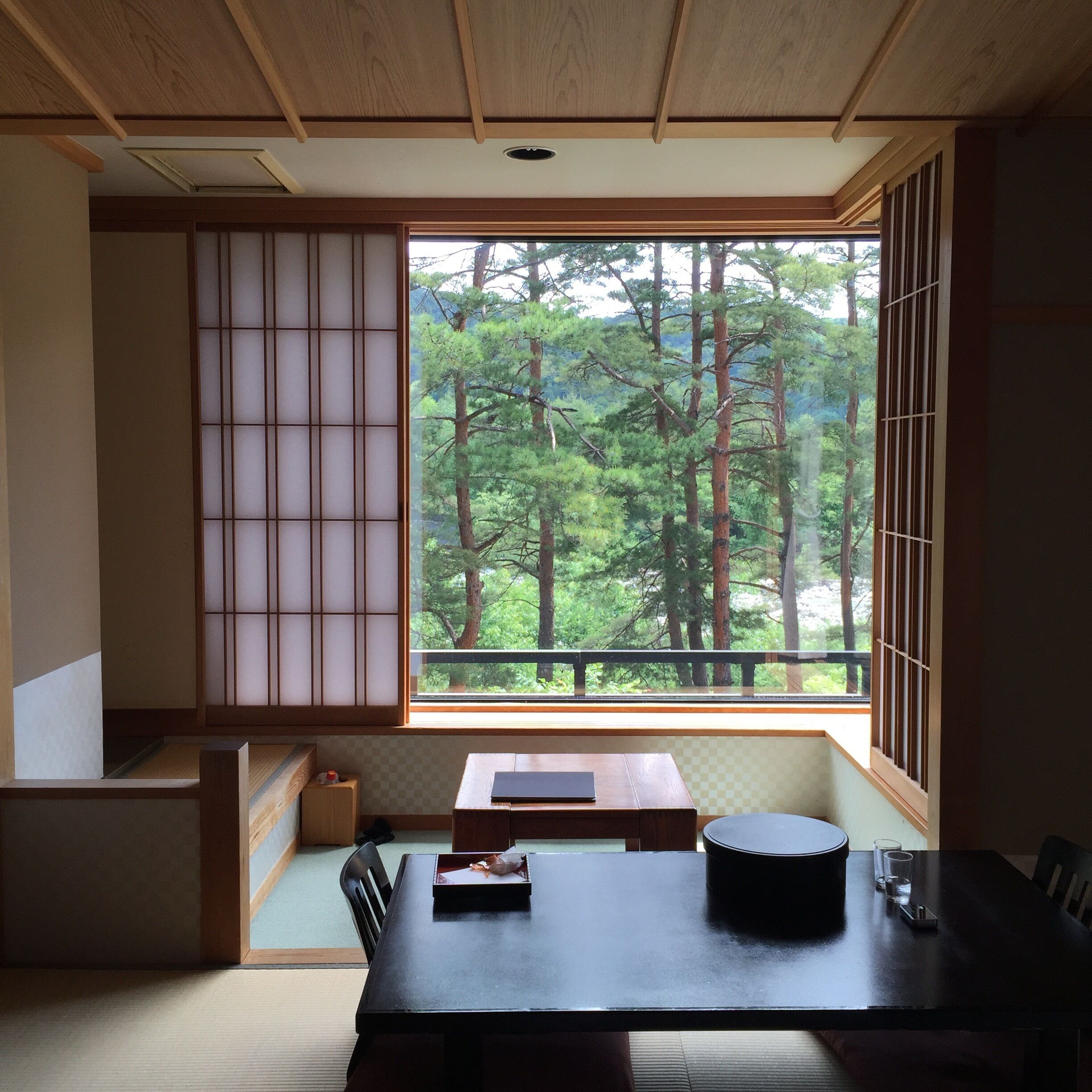 A traditional Japanese room with tatami flooring, a low table, and a large window overlooking a forest.