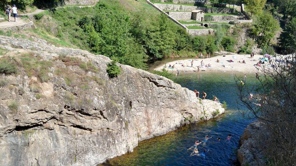 A rocky riverbank with people swimming and sunbathing on a nearby beach.