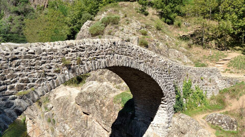 An ancient stone bridge arching over a narrow canyon with rugged terrain and dense greenery on either side.