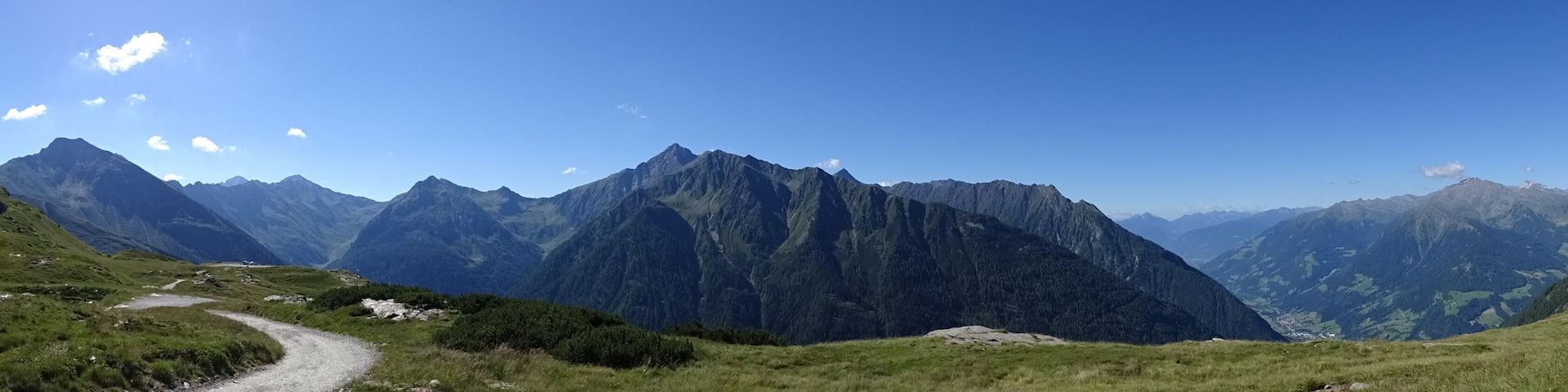 A mountain landscape with a winding path, lush green hills, and distant snow-capped peaks under a clear blue sky.