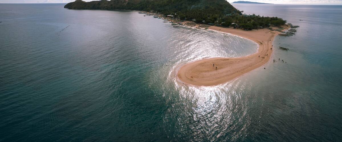An aerial view of a coastal island with a curved sandy beach, clear blue waters, and a backdrop of lush green hills.