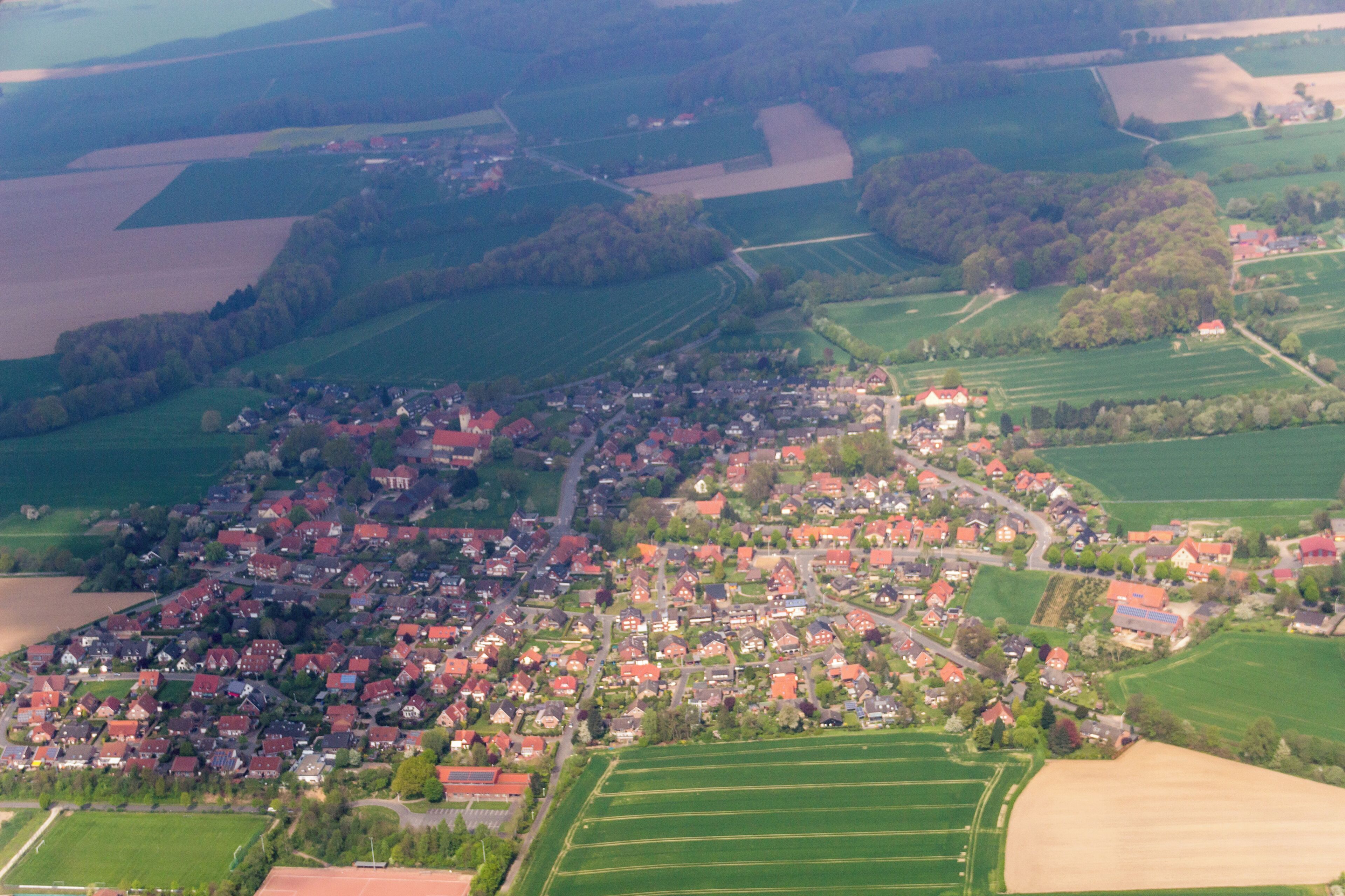 An aerial view of a rural village with red-roofed houses, green fields, and scattered trees.