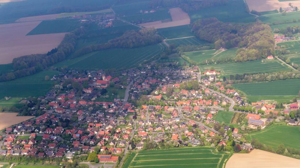 An aerial view of a rural village with red-roofed houses, green fields, and scattered trees.