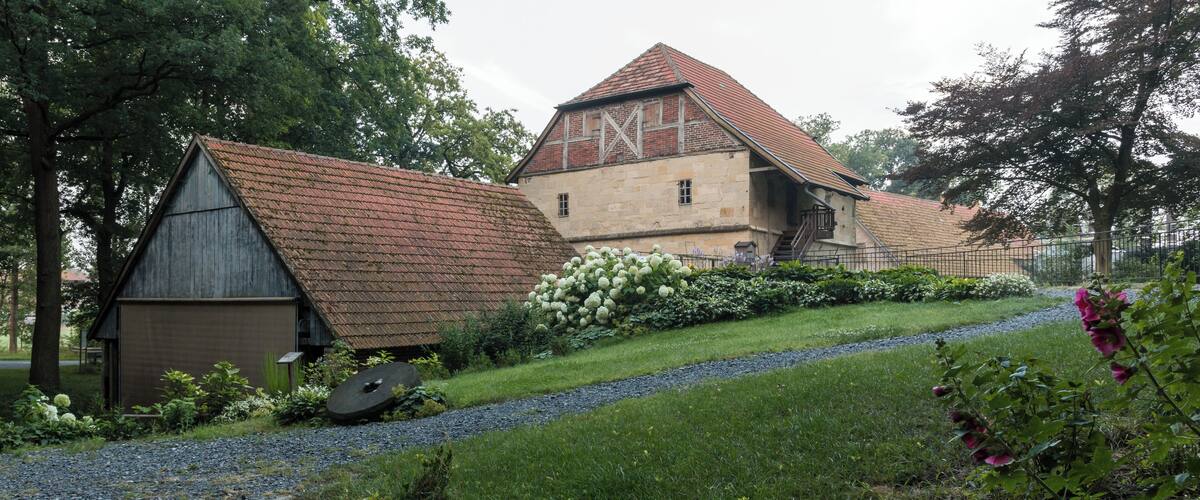 A traditional stone building with a red-tiled roof, surrounded by lush greenery and a gravel path.
