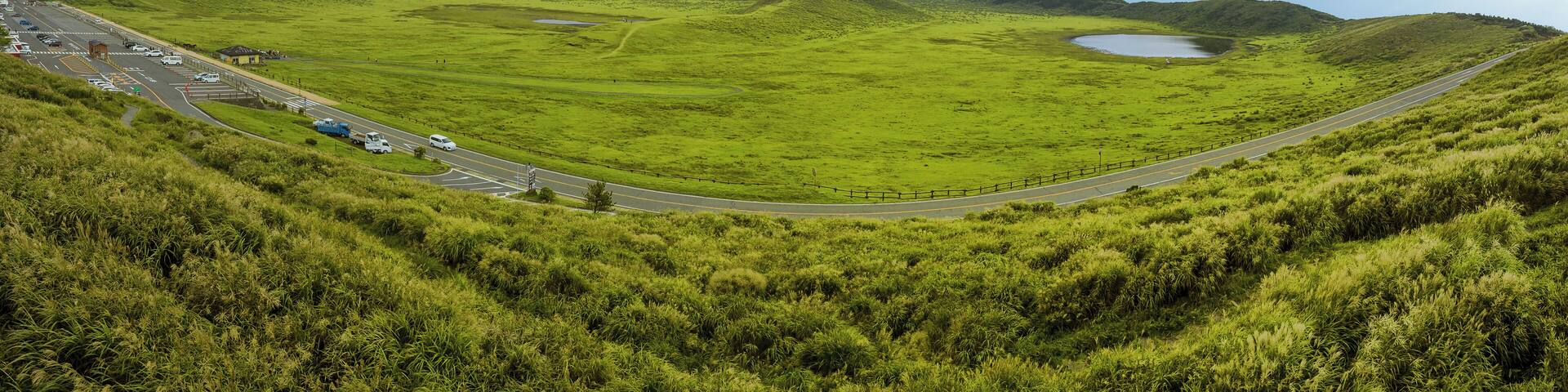 A winding road through a lush green valley with a lake and a mountain in the background.