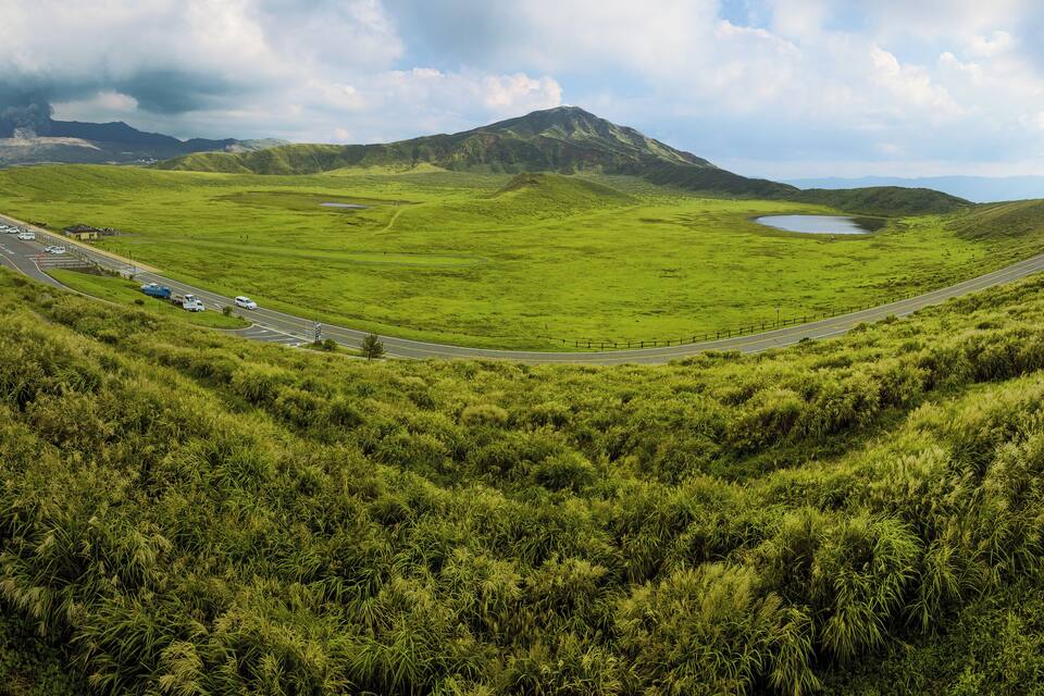 A winding road through a lush green valley with a lake and a mountain in the background.