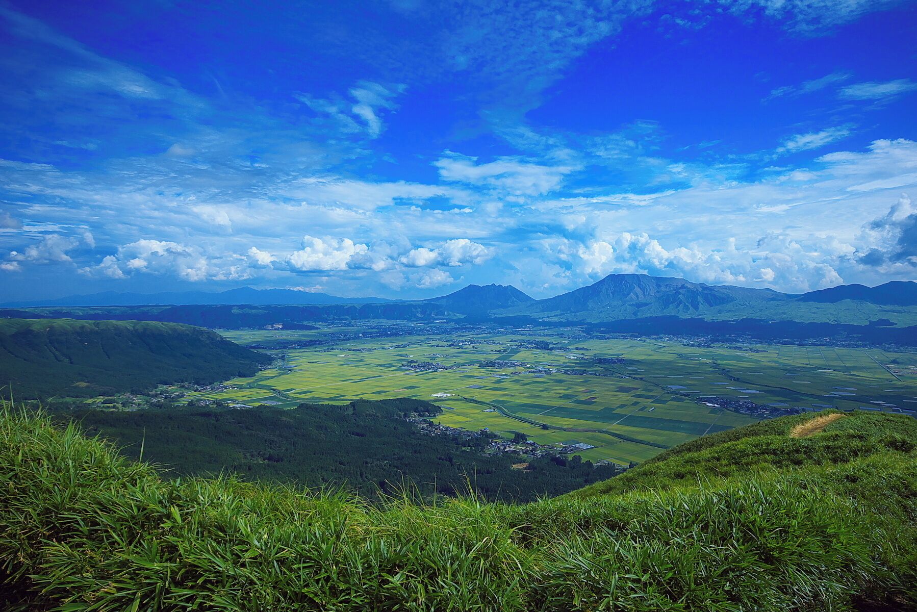 A panoramic view of a valley with green fields, scattered houses, and mountains in the background under a partly cloudy sky.