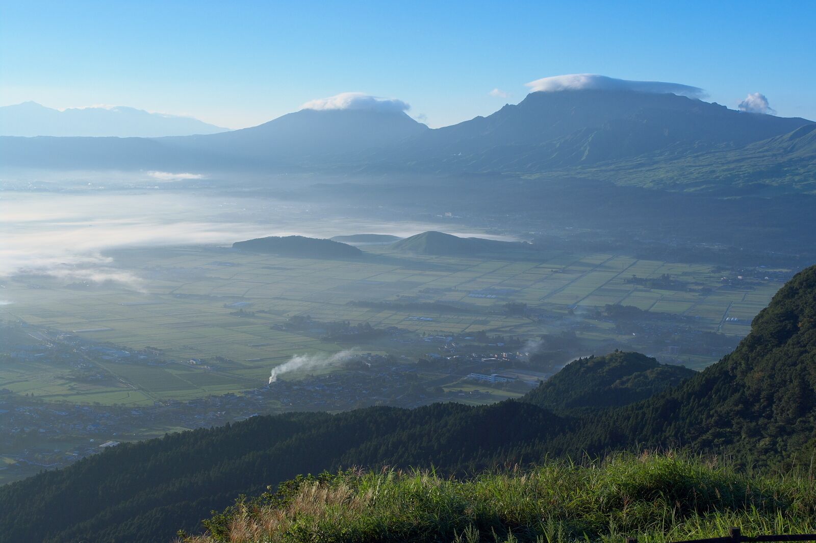 A mountainous landscape with a valley, fields, and a distant mountain range.