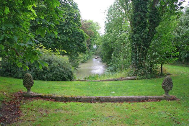 A tranquil garden with a pond, lush greenery, and two decorative urns.