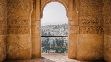 Uma porta arqueada com esculturas detalhadas, dando acesso a uma vista da paisagem urbana com edifícios e árvores.