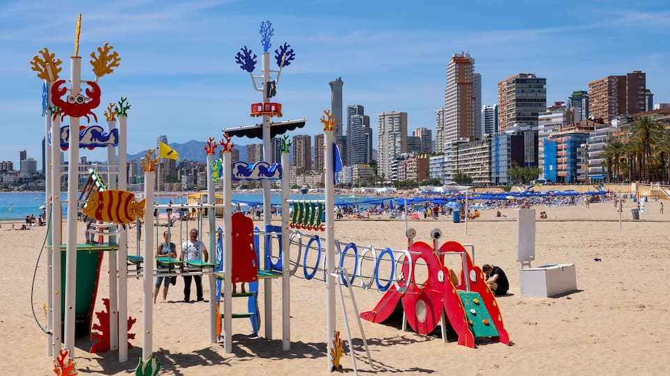 A beach playground with slides and climbing structures, a sandy beach, and a city skyline with tall buildings in the background.