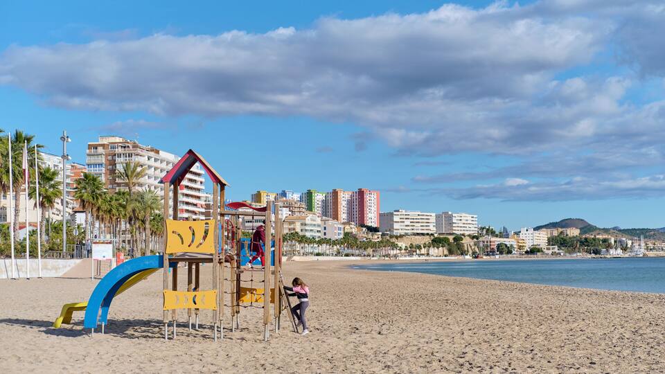 A beach with a playground, palm trees, and a cityscape in the background.