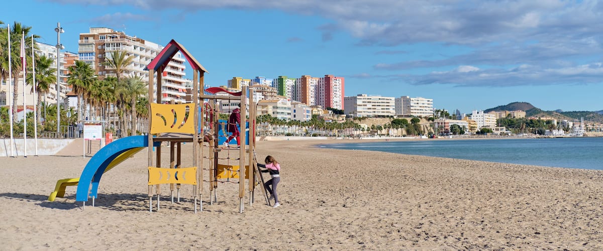 A beach with a playground, palm trees, and a cityscape in the background.