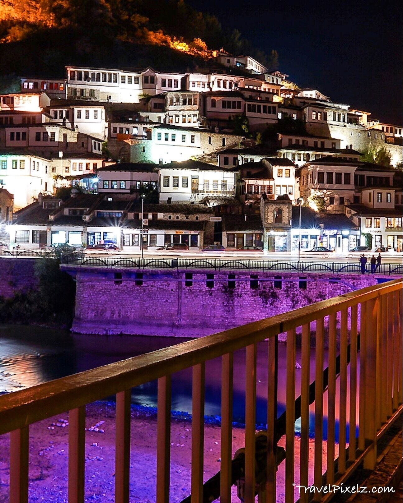 A night view of a terraced hillside with illuminated buildings and a stone bridge over a river.